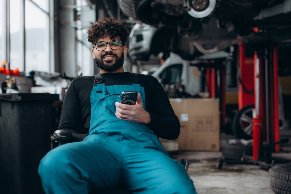 Mobile mechanic in uniform smiling while holding a phone in an auto repair shop